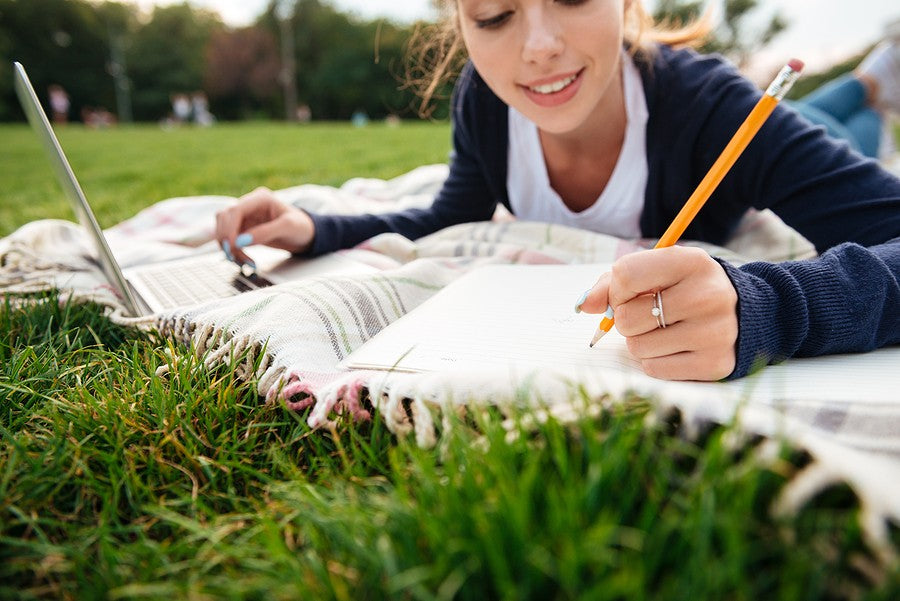 A homeschooled student writing in a notebook while studying outdoors on a blanket.